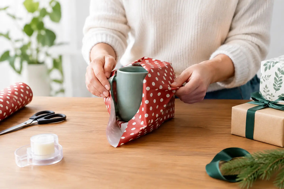 Hands wrapping a green mug in red spotted paper beside scissors, tape and ribboned gifts